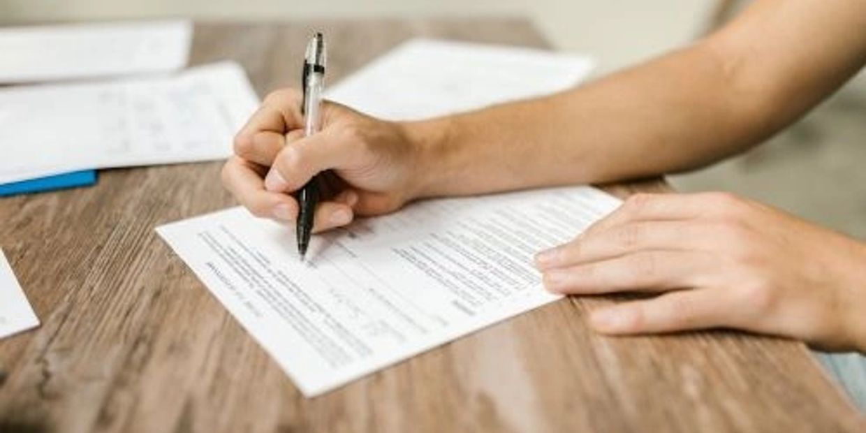 Person writing on a document with a pen at a wooden table.