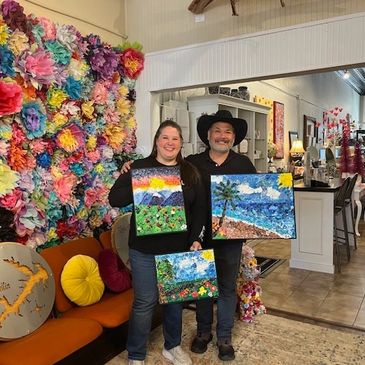 Two people proudly display colorful landscape paintings in a cozy, art-filled room.
