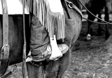 Black and white photograph of a rider’s boot resting in a stirrup on horseback.