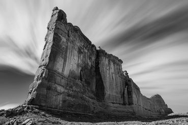 Black and white photograph of a tall rock formation rising against a streaked sky.