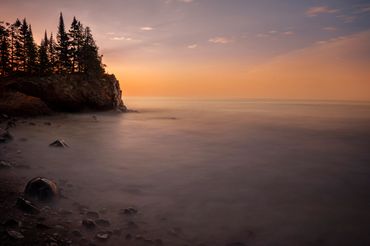 Color photograph of a quiet coastal shoreline at sunset with trees along the cliff edge.