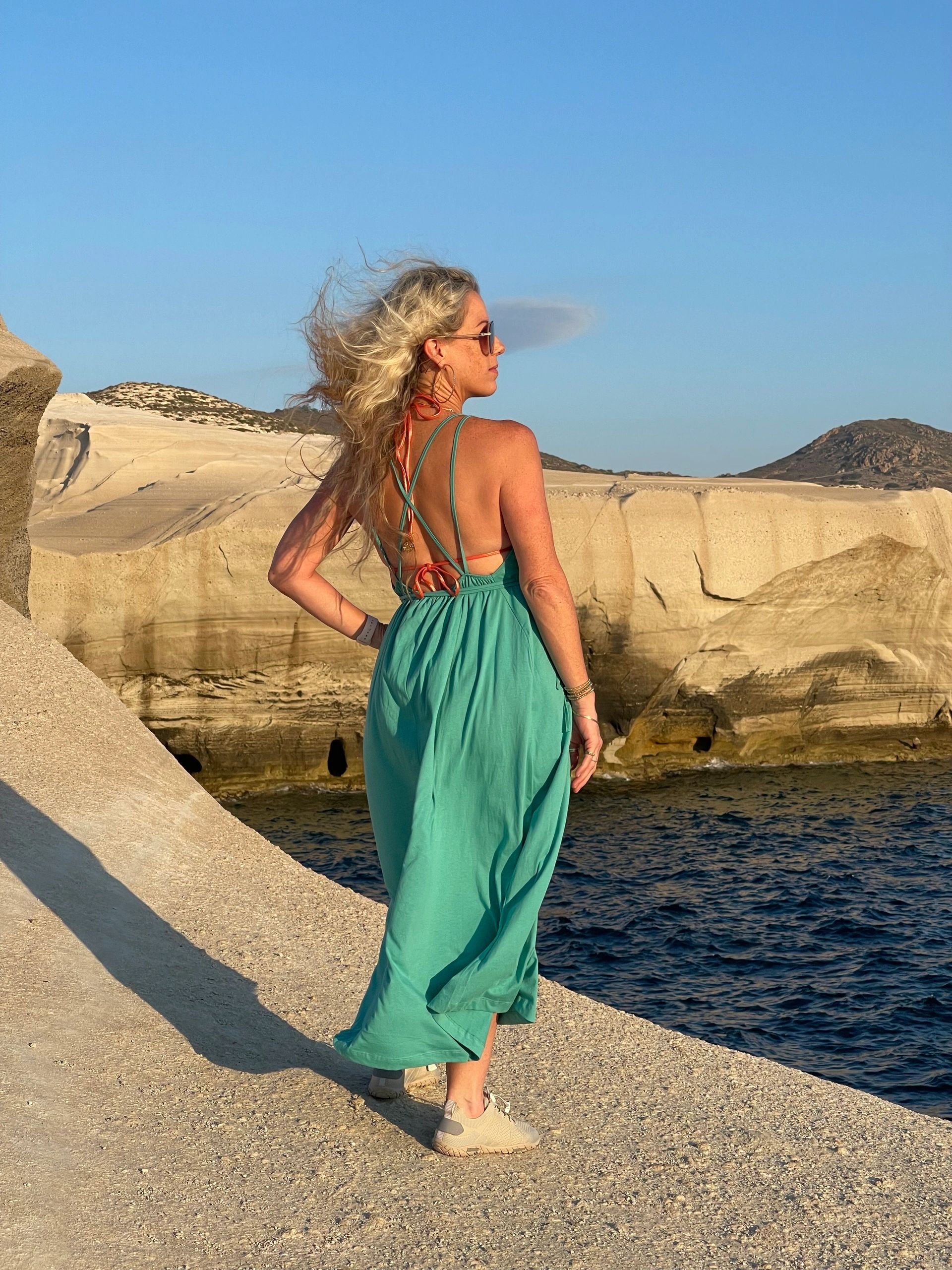 Woman in teal dress stands on rocky shore with ocean and hills in background.