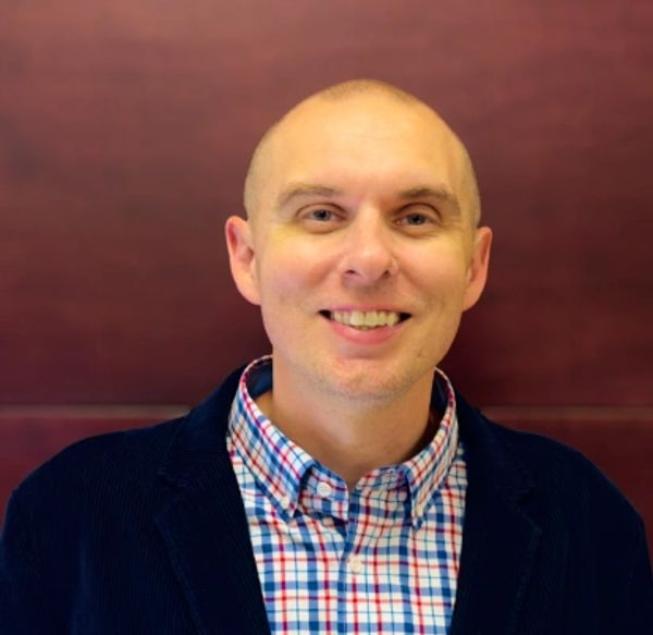 Smiling man in a checkered shirt and dark jacket against brown background.