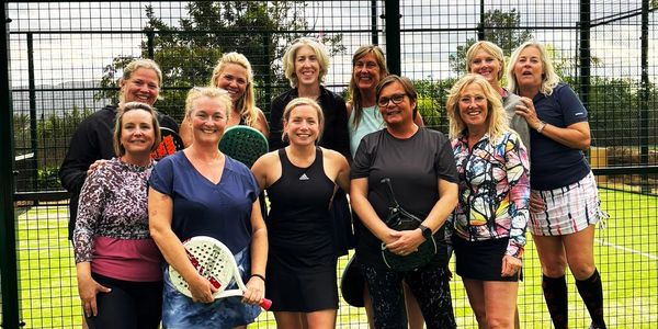 Group of women posing on a paddle tennis court with rackets.