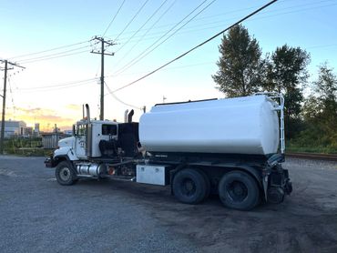 White tanker truck parked on gravel near power lines and trees at sunset.