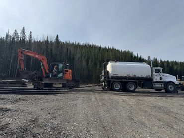Excavator and tanker truck on a railway track in a forested area.