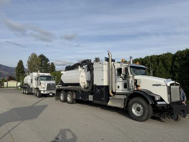 Two large white tanker trucks parked on a road with trees and hills in the background.