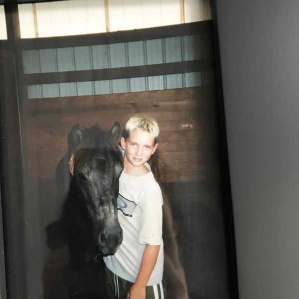 A boy with blonde hair hugging a black horse indoors.