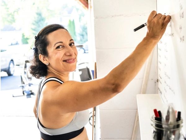 Smiling woman in gym clothes writing on a whiteboard in a bright workout space.