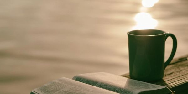 A book and cup on a wooden surface by water during sunset.