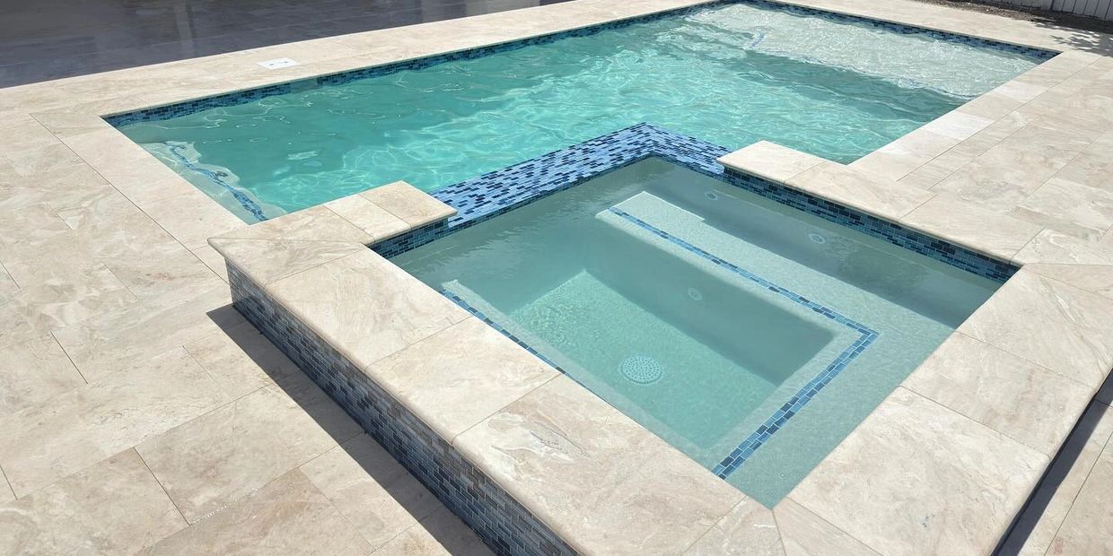 A modern pool with an attached spa, featuring light stone tiles and blue mosaic accents.