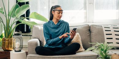 Woman relaxing on sofa using smartphone with smart glasses and earbuds.