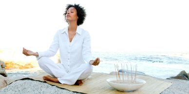 Woman in white meditating peacefully by the ocean on a mat.
