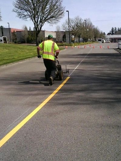 Worker in a high-visibility vest painting a yellow centerline on a road using a striping machine.