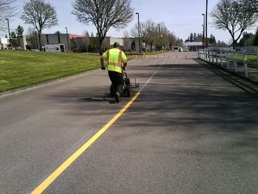 Worker in a high-visibility vest painting a yellow centerline on a road using a striping machine.