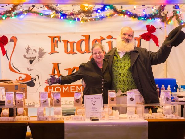 Two cheerful vendors proudly display homemade fudge at a decorated holiday market booth.