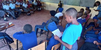 Students gathered in a classroom, one reading aloud to peers seated around.