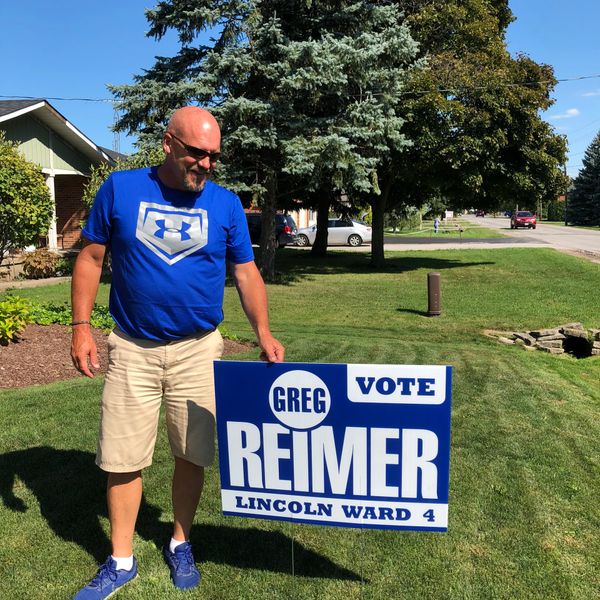 Man in blue shirt posing with a 'Vote Greg Reimer' campaign sign on a lawn.