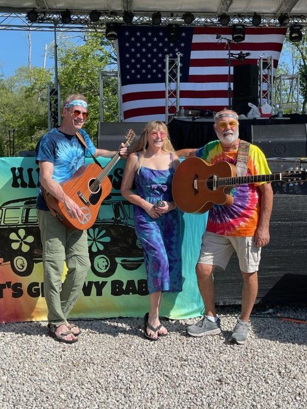 Three people in colorful tie-dye outfits holding guitars, smiling outdoors.
