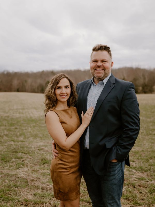 A smiling couple poses outdoors in casual formal attire on a cloudy day.