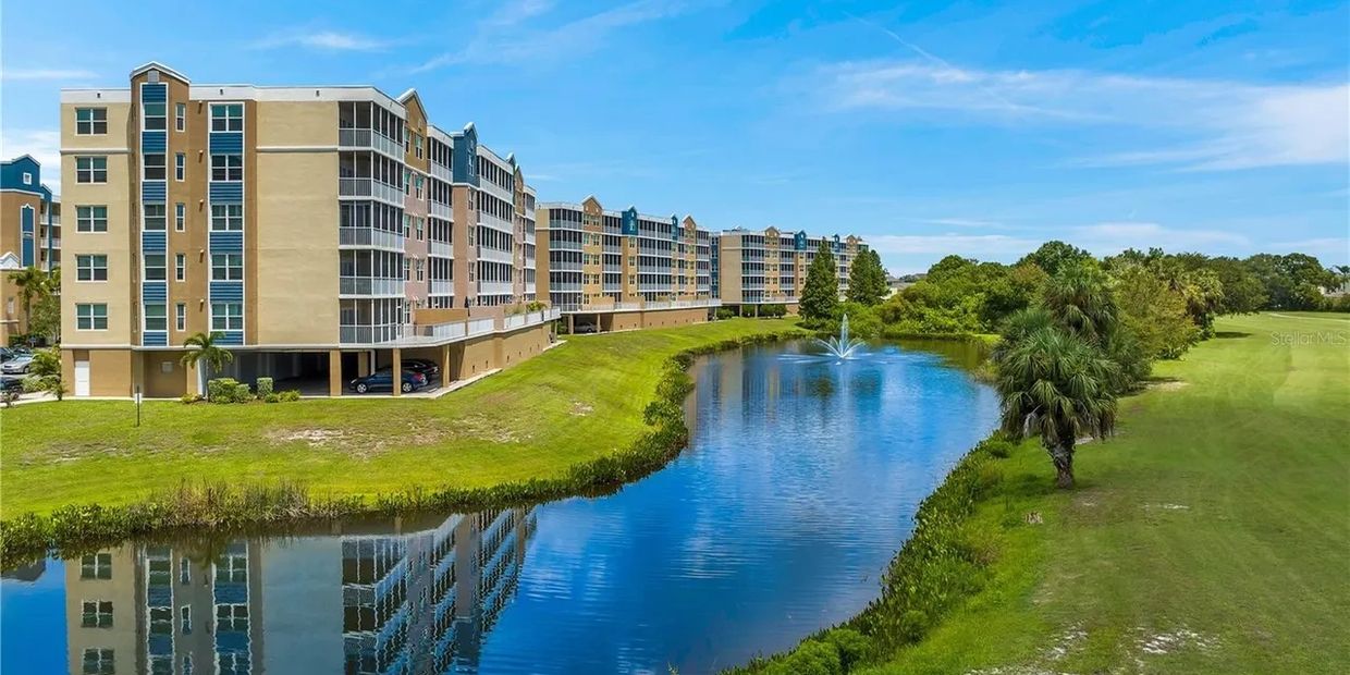 Apartment complex beside a pond with a fountain under a blue sky.