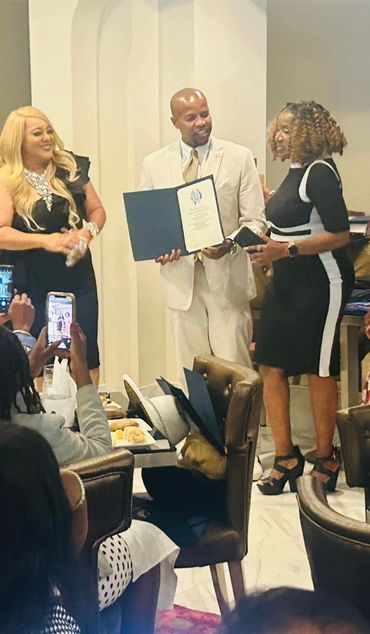 Man in beige suit holding certificate with two women at a formal event.