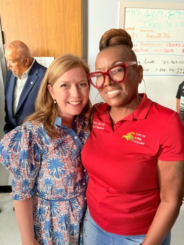 Two women smiling together, one in a patterned dress and the other in a red City of Iowa Colony shirt.