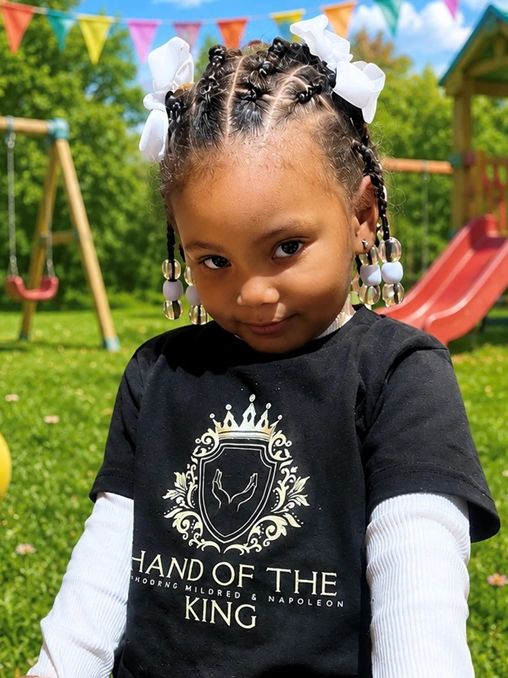 A young girl with braided hair and white bows smiles outdoors in a playground.