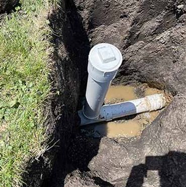 A vertical PVC pipe connected to a horizontal pipe in a muddy trench.