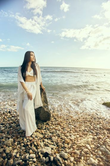 Woman in white outfit standing on a rocky beach by the ocean at sunset.