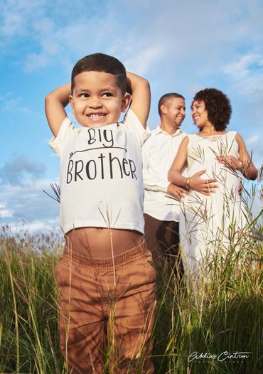 Smiling boy in 'Big Brother' shirt with happy parents and pregnant mother in the background.