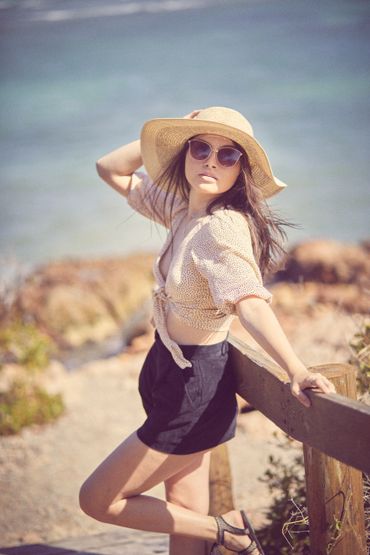 A woman in sunglasses and a sunhat poses by a wooden railing near the beach.