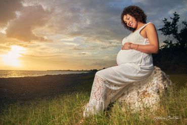 Pregnant woman in white dress sitting on a rock at sunset by the sea.