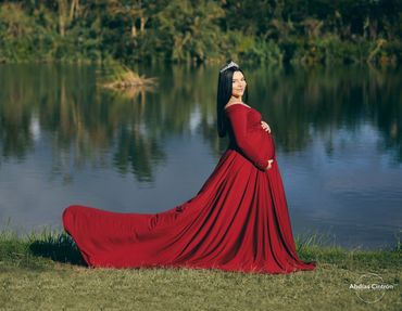 Pregnant woman in a flowing red gown by a serene lake, wearing a tiara.