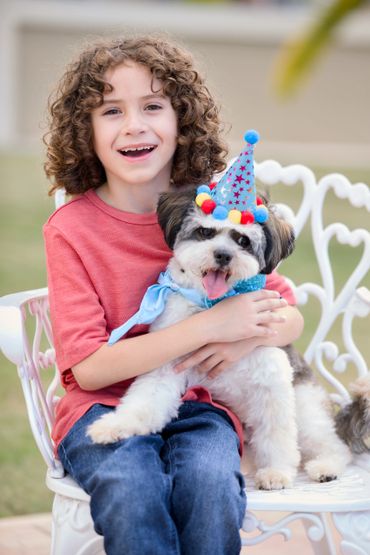 A happy child in a red shirt hugs a dog wearing a festive party hat and blue scarf.