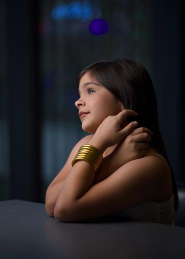 A young girl with gold bangles gazes thoughtfully to the side in a dim setting.