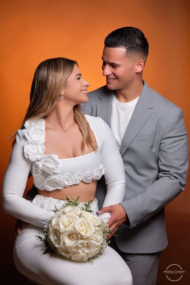 Couple in elegant attire smiling at each other against an orange background.