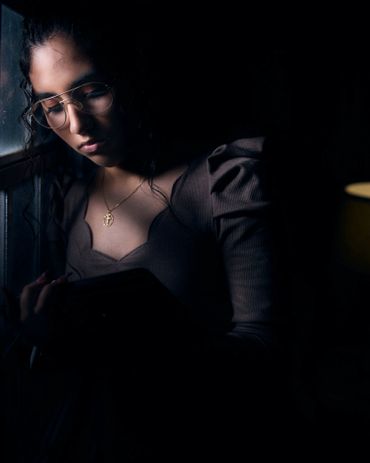 A woman with glasses reads by a window in dim light.