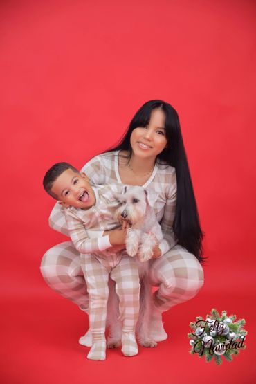 Mother and son in matching pajamas with their dog against a red Christmas backdrop.