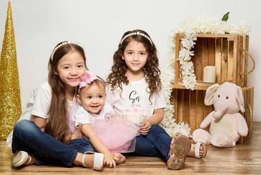 Three young girls sitting on the floor, smiling, with a stuffed elephant and wooden crates decorated with white flowers.