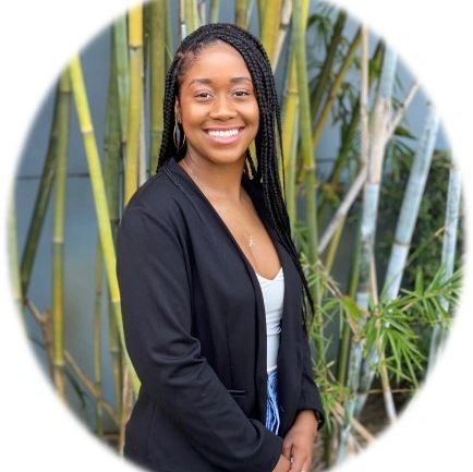Smiling woman with braided hair in black blazer standing by bamboo plants.