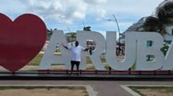 Person posing with large "I ❤️ Aruba" sign under a partly cloudy sky.
