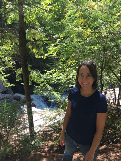 The artist Andrea Sauchelli is standing above the rushing river in the Adirondacks on a sunny day.