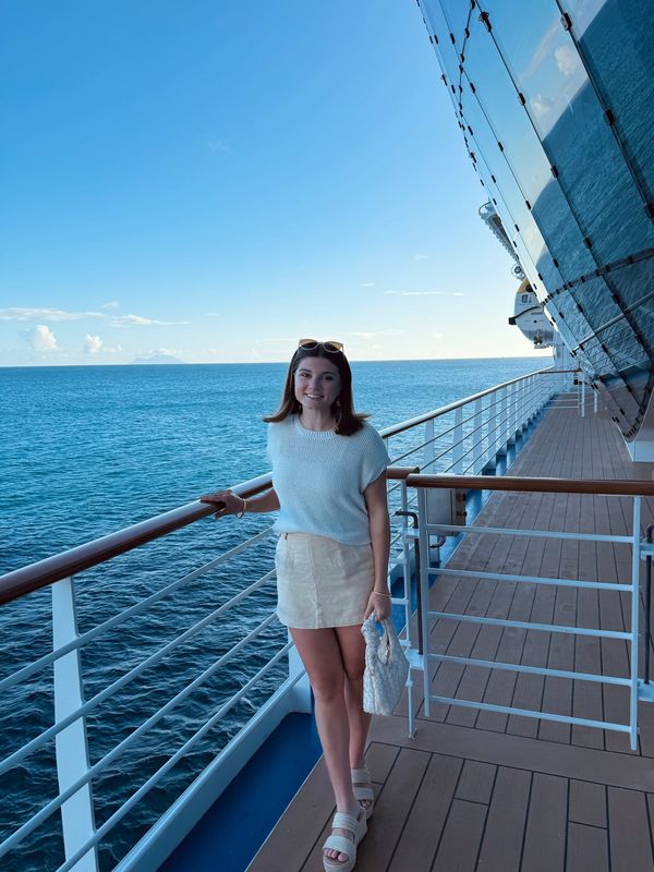 A young woman stands on a cruise ship deck by the ocean, smiling at the camera.