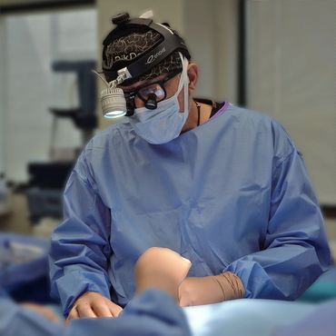 Surgeon in scrubs performing surgery with magnifying glasses and headlamp.