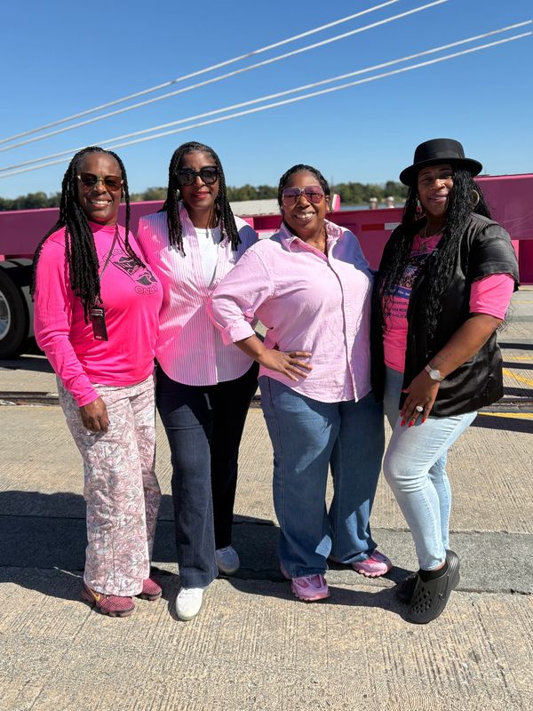Four women smiling outdoors on a sunny day, dressed casually with pink accents.