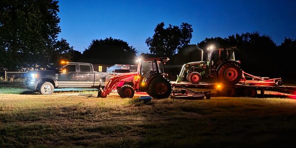 A truck and two tractors with lights on at night in a grassy area.