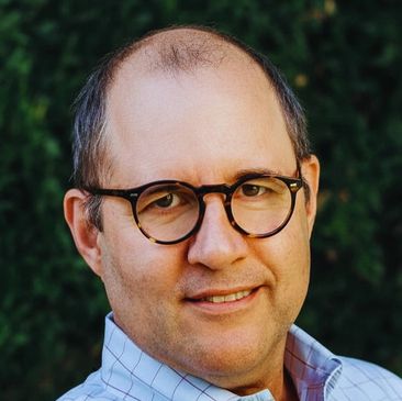 Middle-aged man wearing glasses and a checkered shirt smiling outdoors.