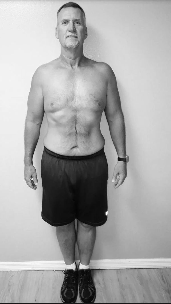 Man standing shirtless in black shorts and sneakers against a plain wall.