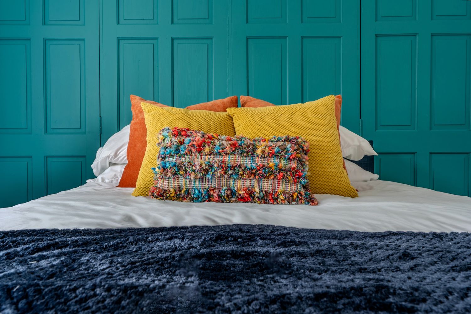 Colorful pillows arranged on a white bed against teal paneled wall.